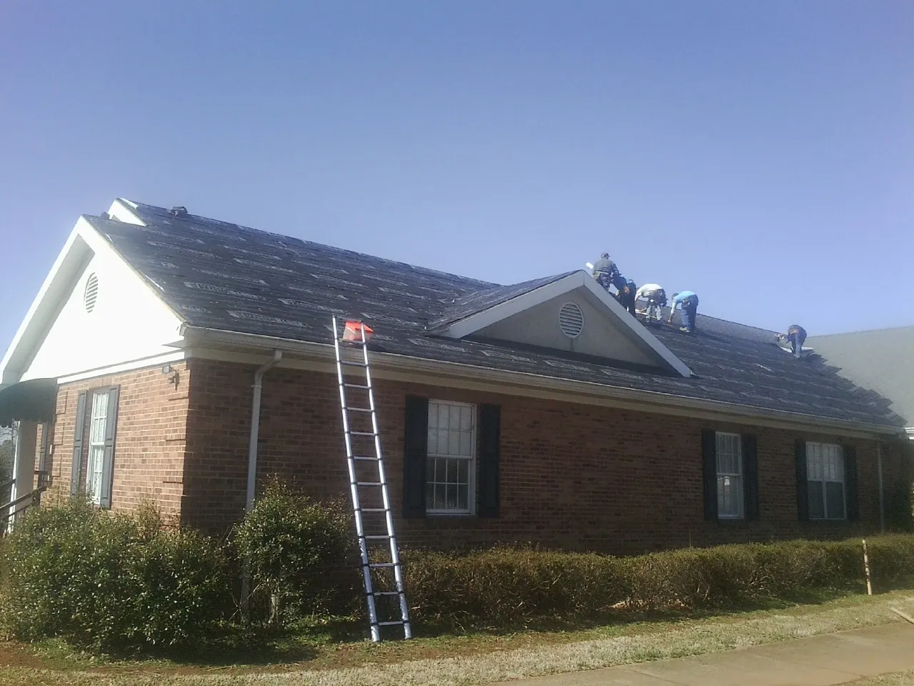 Crew tearing shingles off a brick ranch roof with a ladder set — Metal Roof Repair work by our crew in Benedict